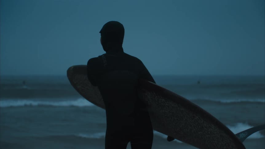 Surfer wearing a full body wet suit holds a surfboard in front of the ocean at evening