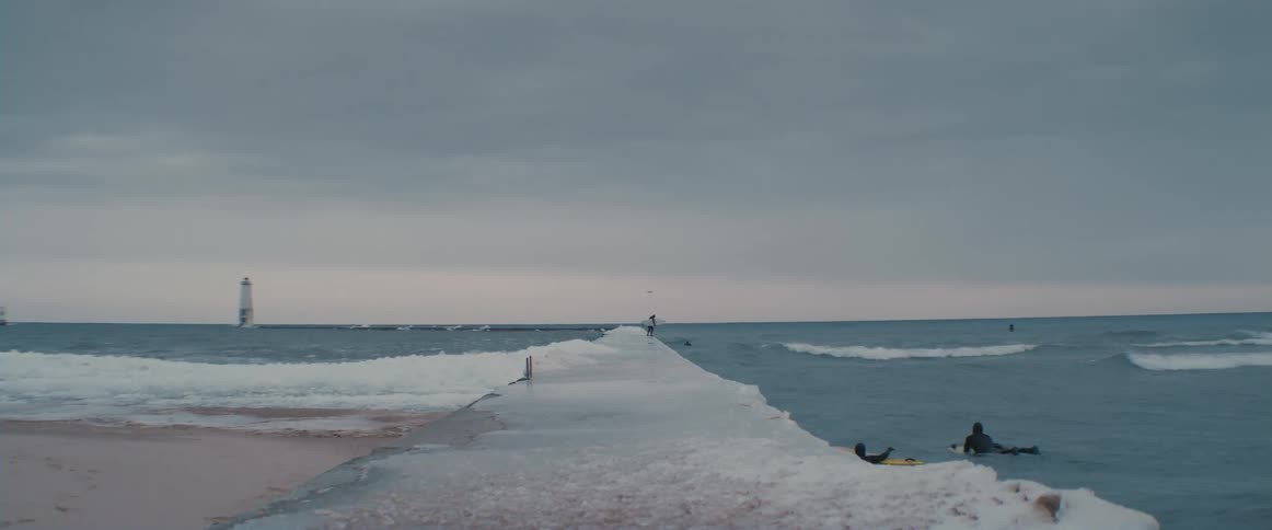 A surfer jumps off a jetty into the cold water with his board while others paddle towards him