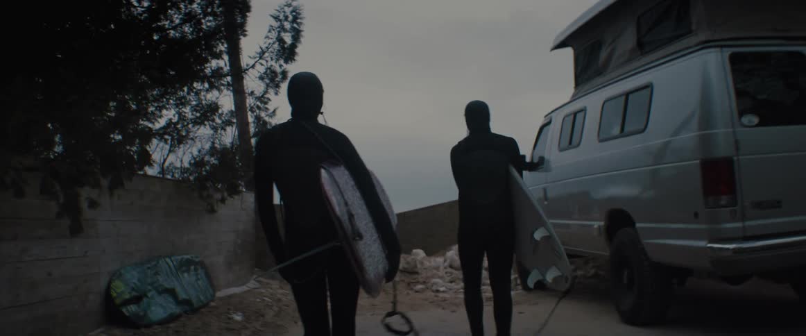 Two surfers wearing full body wet suits pick up surfboards from the floor near a parked van