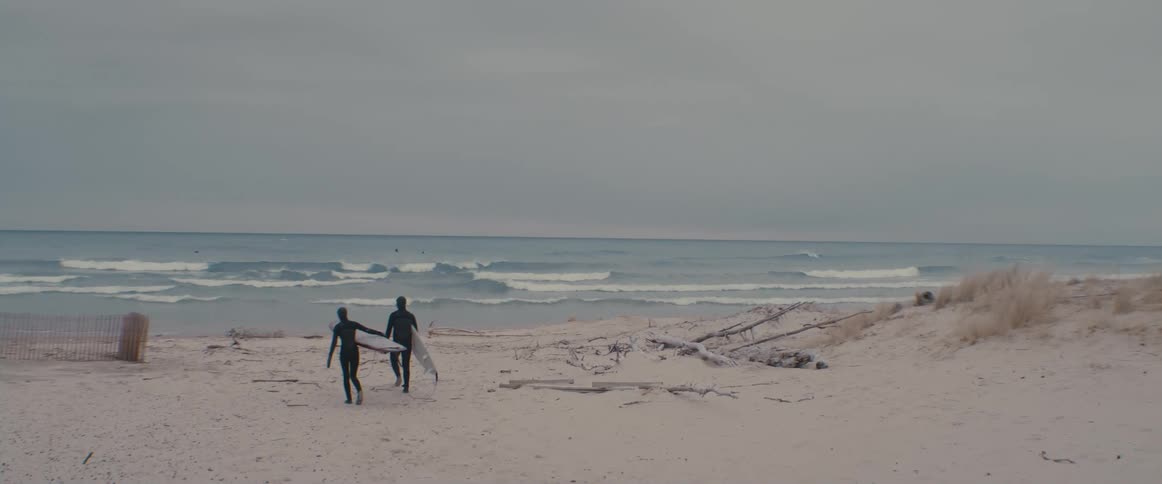 A man and woman wearing wet suits carry their surfboards through the sand towards the waves
