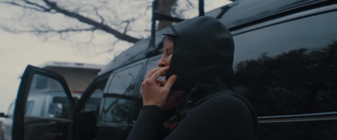 Woman struggles to put a wet suit hood over her head next to a black van
