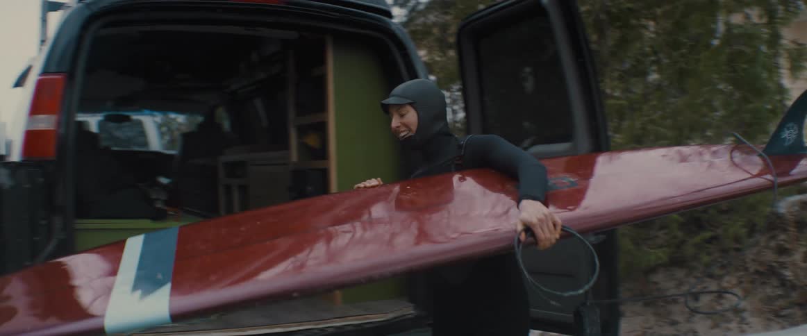 A woman smiles as she pulls a surfboard out of the van and carries it