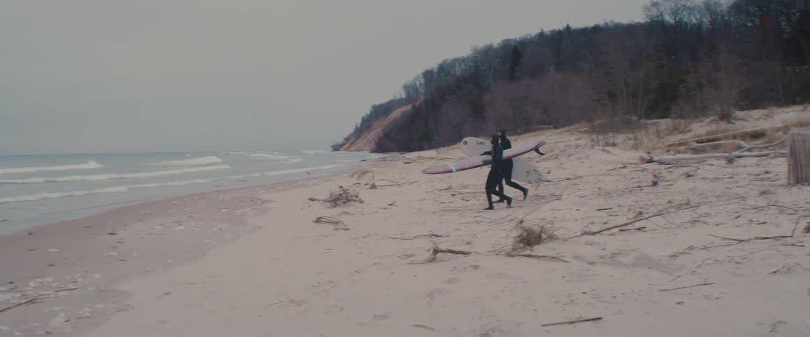 Surfers clad head to toe in wet suits carry surfboards across beach