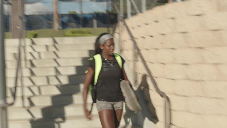 A woman carrying some equipment walks over to a bench and sits down