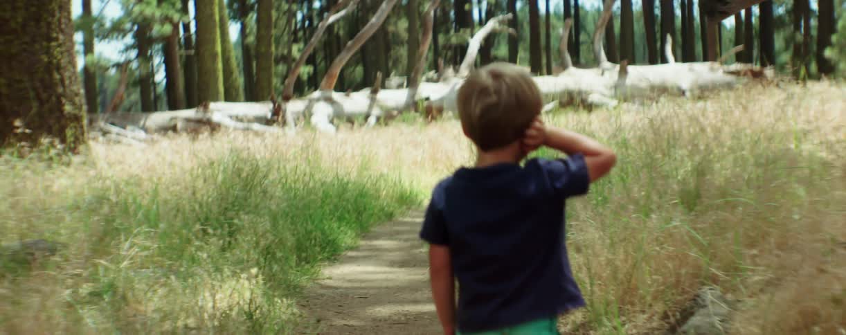 A toddler boy with his hands on his ears walks on a dirt path in a wooded area
