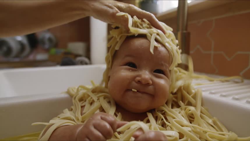 A toddler is sitting in a kitchen sink covered in wet noodles while nibbling on them and the mother continues to playfully add more