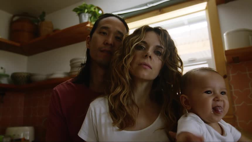 A woman holds her baby while posing with her partner in their kitchen