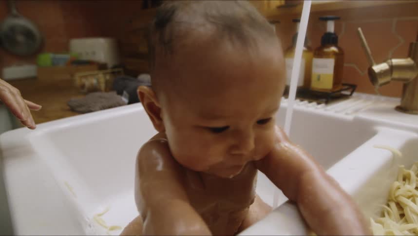 A woman is soaking noodles in a kitchen sink while a toddler is sitting in a secondary sink playing with the wet noodles