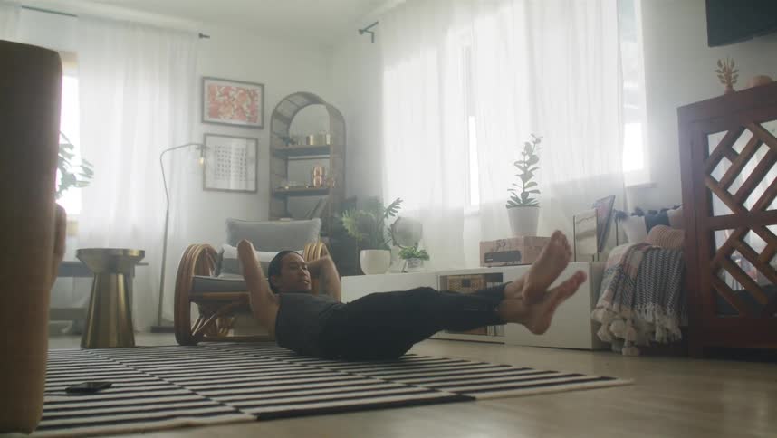 Man with long hair sleeveless shirt and black pants does sit ups in living room at home