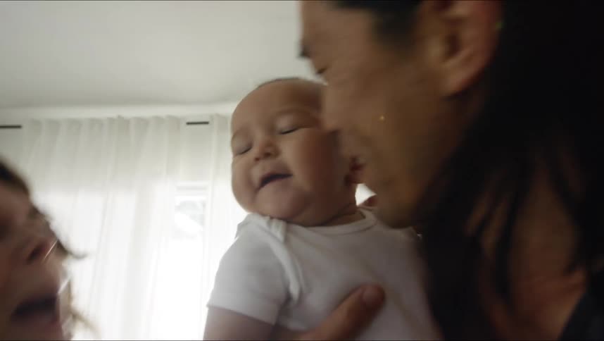 A father is holding his son while both he and his wife kiss the toddlers cheeks at the same time while standing in a living room during the day