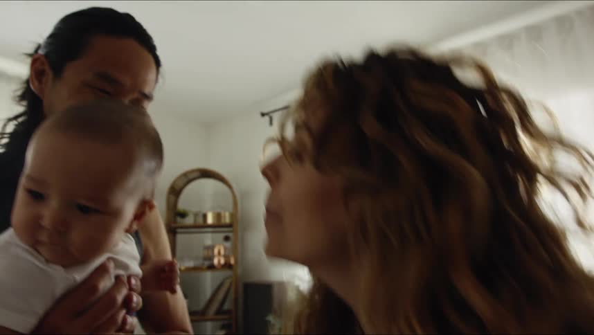 Father with black undershirt and long hair holds baby son and dances with mother with brown wavy hair in living room at home