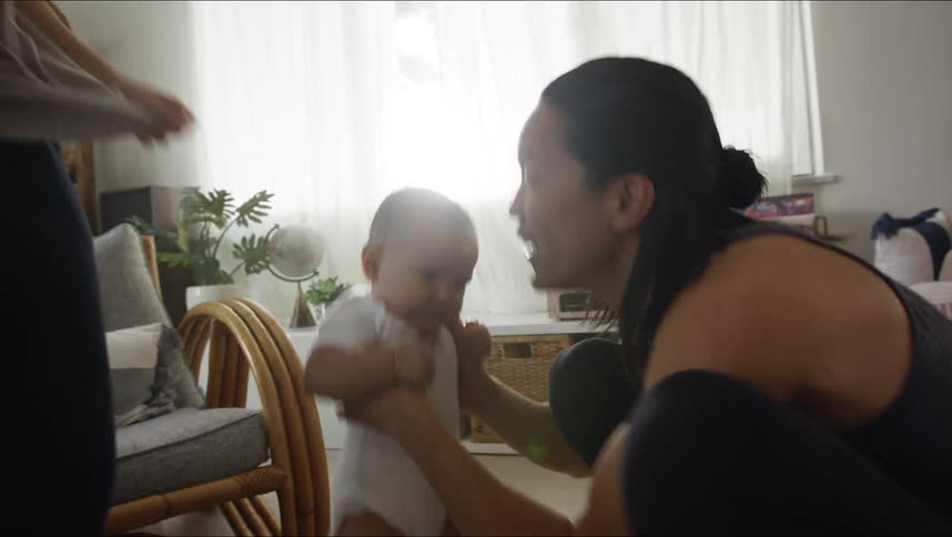 Father with long hair and black sleeveless shirt plays with baby son lifting him up in living room at home