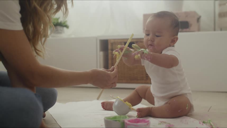 A baby sits on a floor with dishes with a colored substance in them while a woman hands a stick to him and he puts it in his mouth