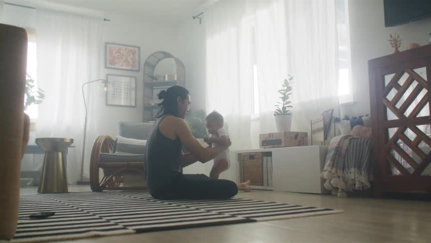 Father with long hair sleeveless shirt and pants plays with baby son in living room at home
