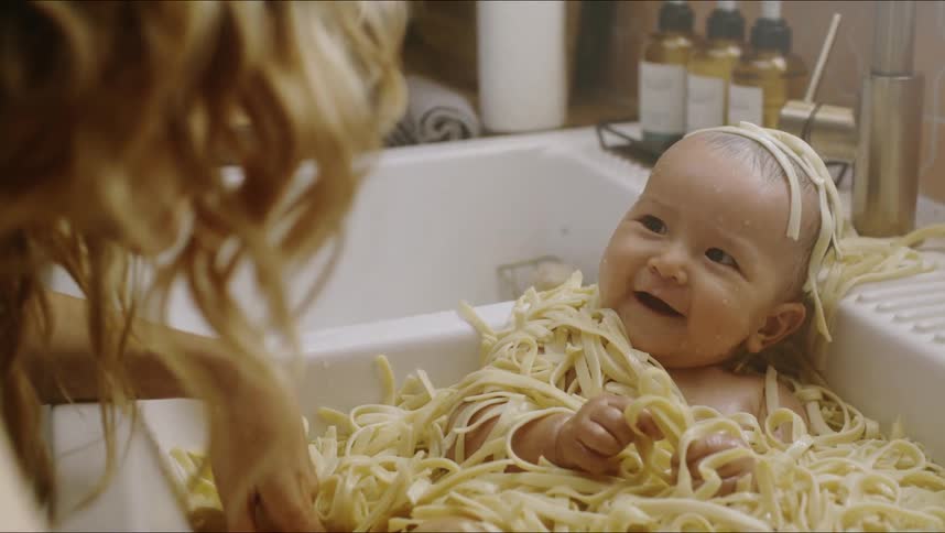 Smiling baby covered in cooked noodles sitting in a kitchen sink