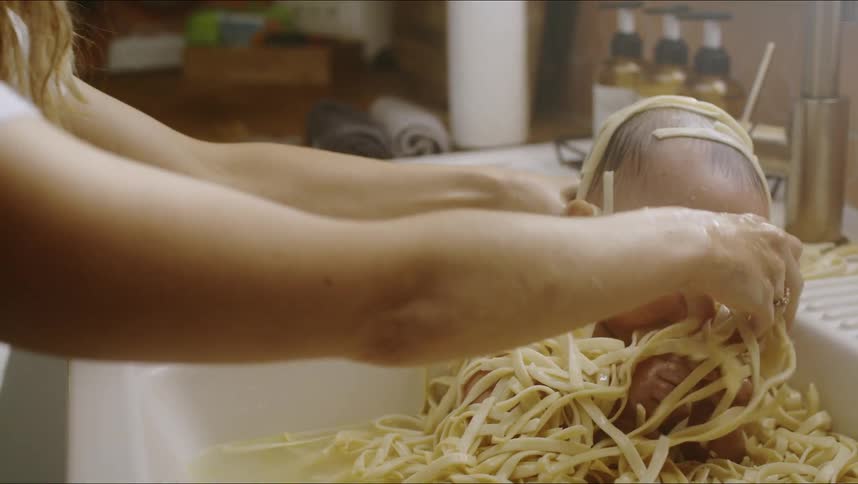Baby sits in kitchen sink surrounded by noodles and eats them as a woman coats the child in noodles