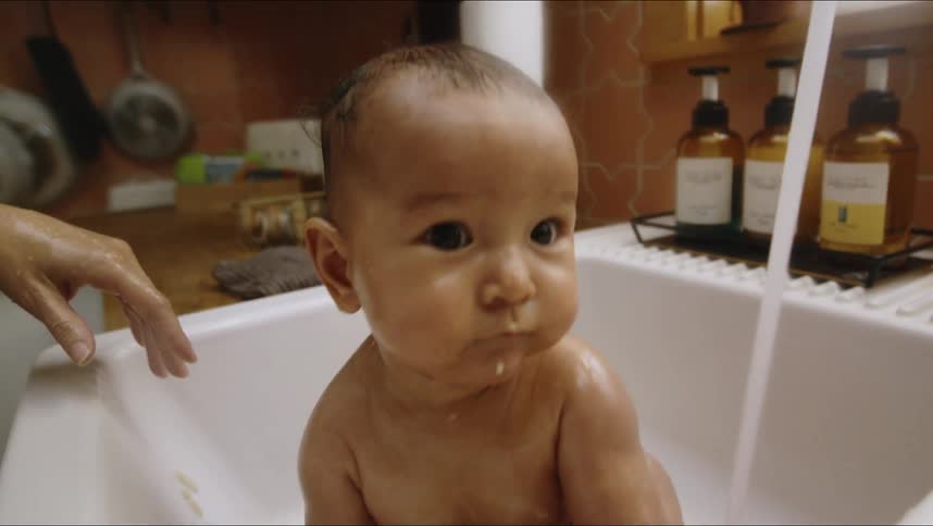 A baby sits in a kitchen sink while the water runs and his mother stands by him