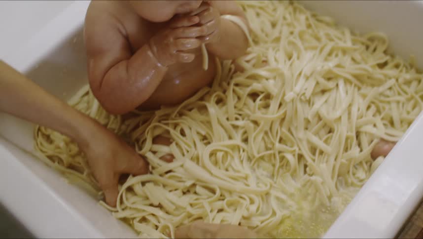 Woman piles more noodles onto baby eating while sitting in a kitchen sink full of noodles