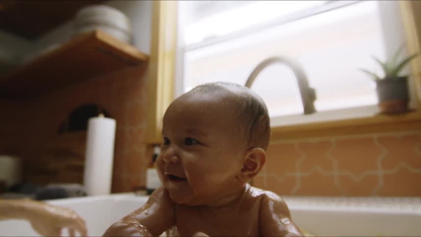 A baby splashes in a kitchen sink filled with noodles while a woman stands by