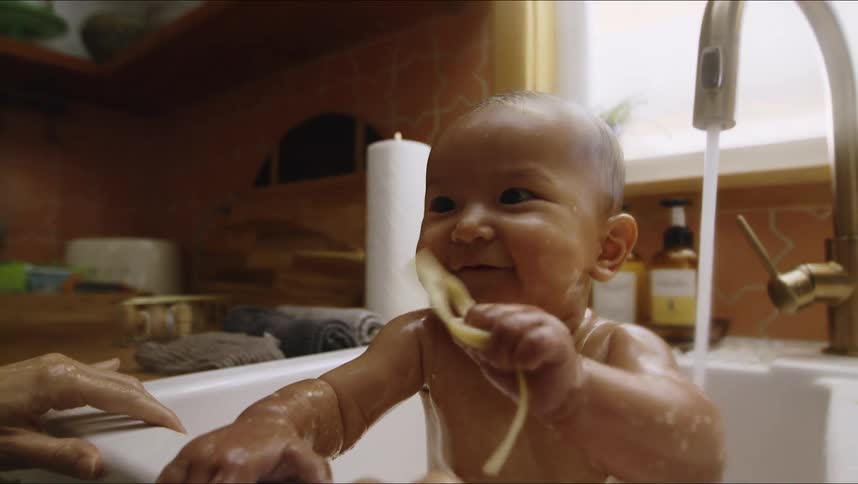 A small infant sits in a kitchen sink playing with a pasta noodle
