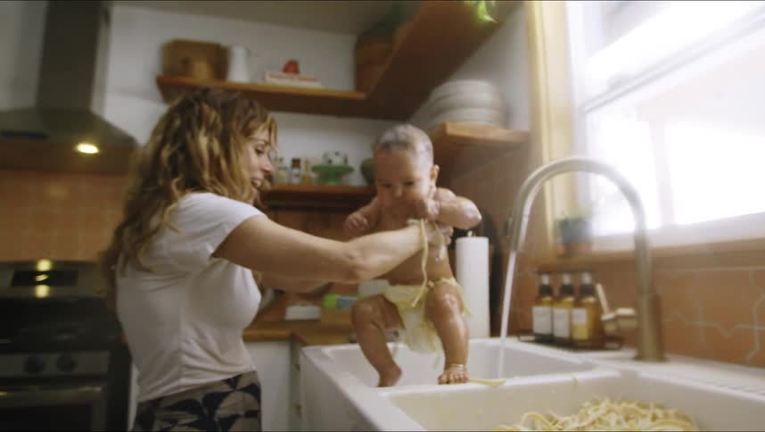 A woman pulls her infant out of a kitchen sink full of pasta