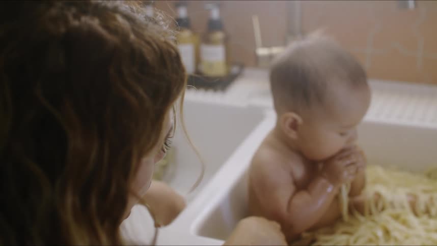 Mother smiles as she watches baby sit in sink full of noodles eat some