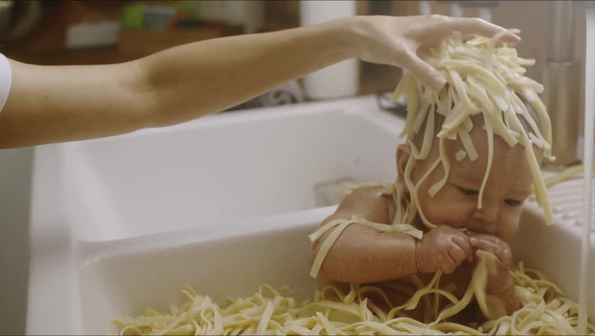A small baby sits in a kitchen sink filled with pasta while the water runs and his mother puts pasta on top of his head and then sits him up when he starts to slip down in the sink