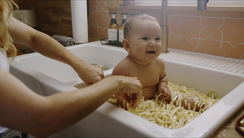 A baby sitting in a kitchen sink full of pasta laughing as mom spreads the noodles all over