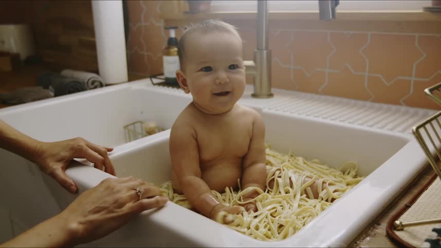 Mom smiles at baby sitting in a sink full of pasta grabbing handful playing and eating