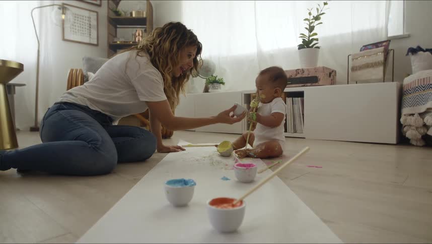 Mother and baby son sitting at the floor painting with paint brushes and water colours while the baby puts a paint brush in the mouth