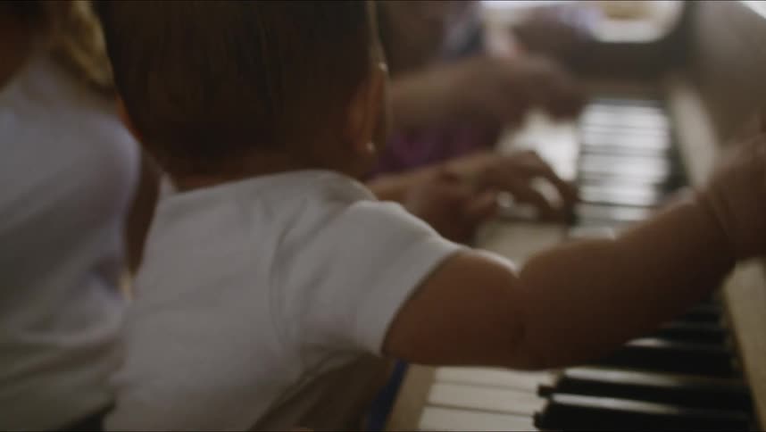 Mother with brown wavy hair and white t shirt holds baby son and plays piano next to father