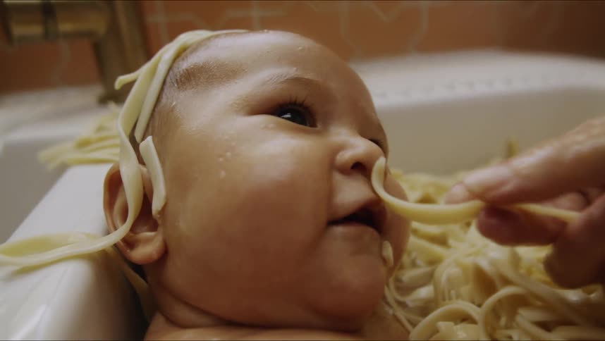 A woman feeds her son pasta while the baby sits in a sink full of fettuccine