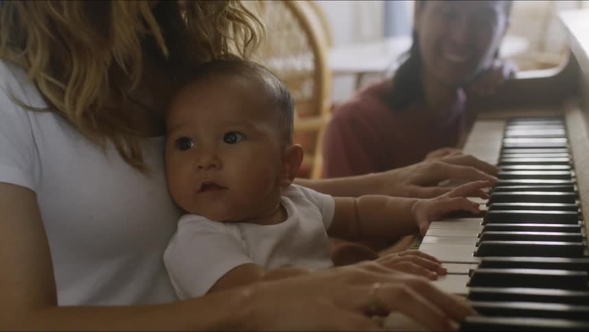 Baby sits on moms lap joyfully banging away at the piano keys as dad looks on