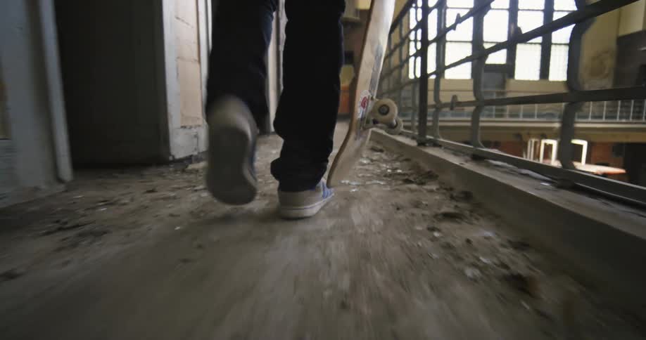 Person carrying skateboard walks along abandoned building walkway near railing passing broken glass shards
