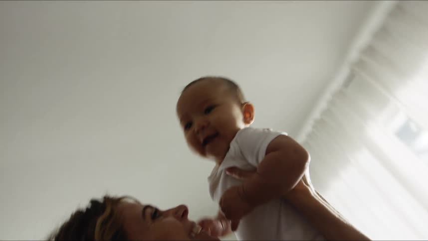 Woman lifts a baby up in the air and back down again while indoors