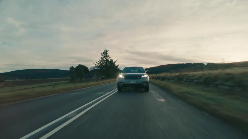Vehicle drives past pastures and buildings with hills in background