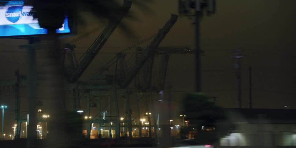 Cars travel on a highway past large cranes lights and a billboard