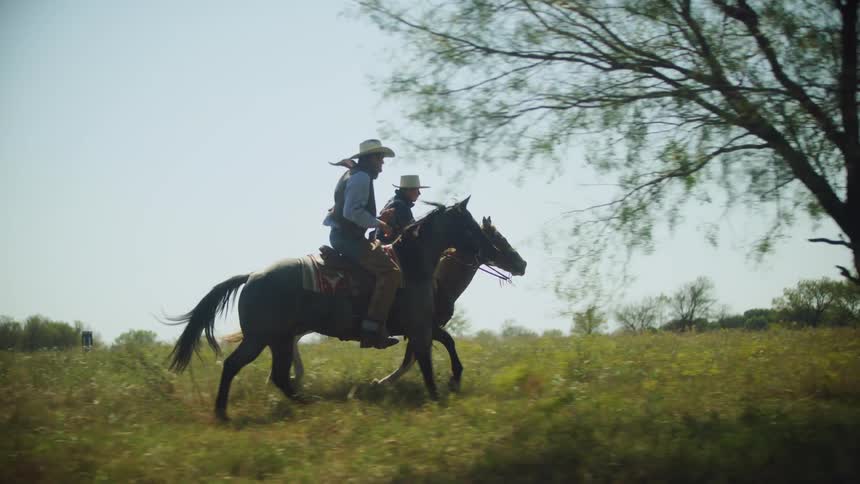 Two cowboys ride horses at a gallop across grass past trees on a sunny day