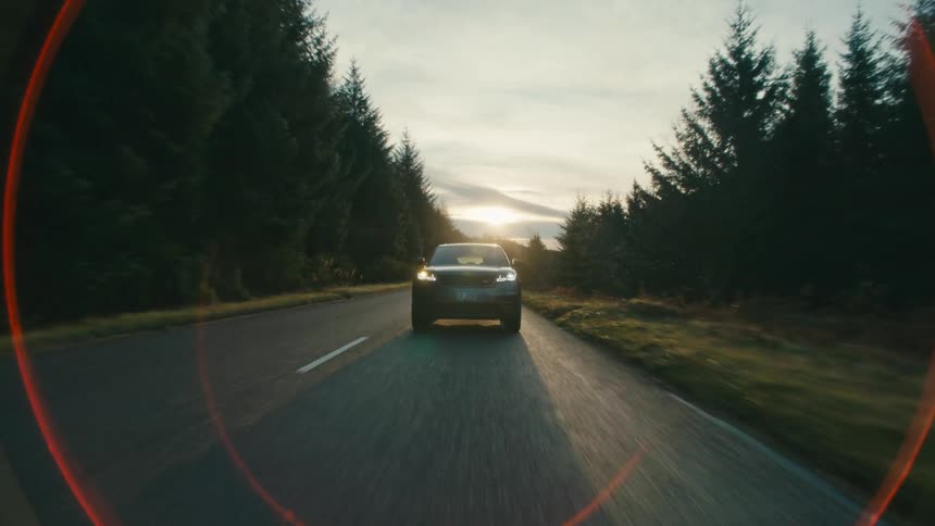 An automobile is traveling on a forest road with the sun on the horizon behind it
