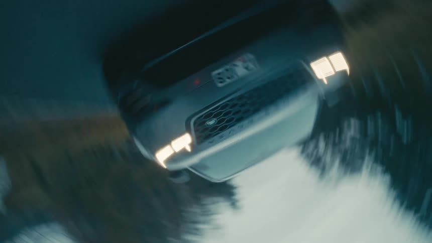 A car is traveling on a forest road past a lake with the headlights on