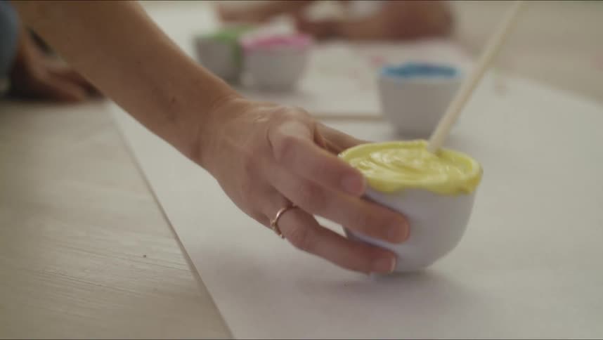 A woman sitting on the floor next to art paper in her home with her toddler child reaches for a small bowl of paint in the daytime