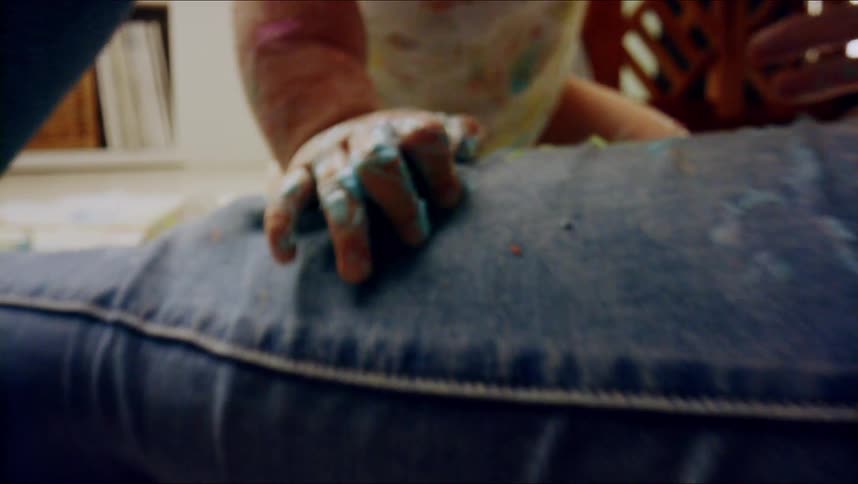 A baby who is covered in food is resting on a womans leg on the floor