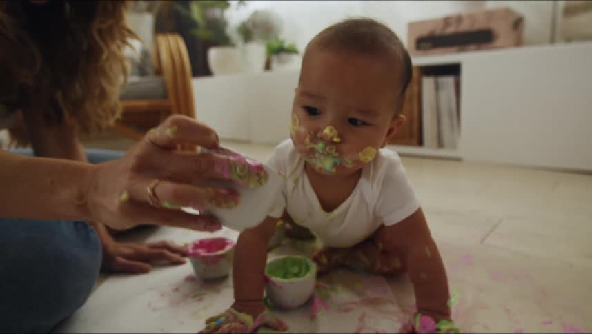 A baby with frosting on his face crawls on the floor as his mother holds up a bowl of frosting