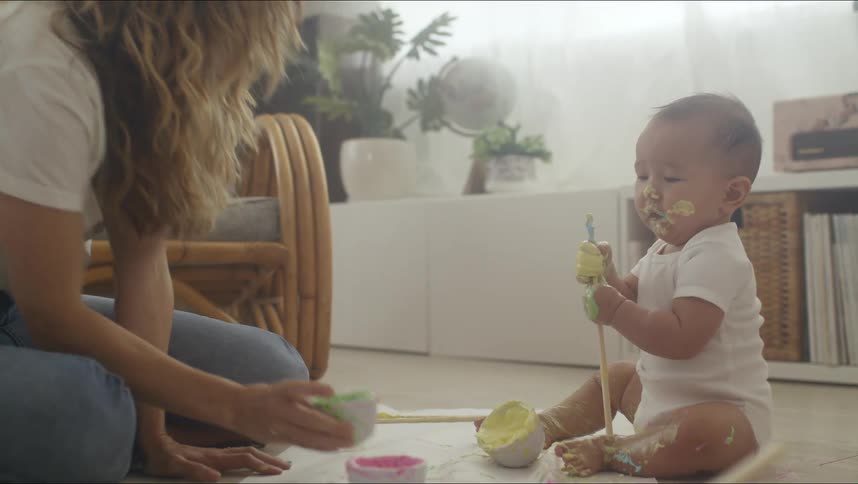 Mother sits with baby in living room as child eats colored pastes off brush
