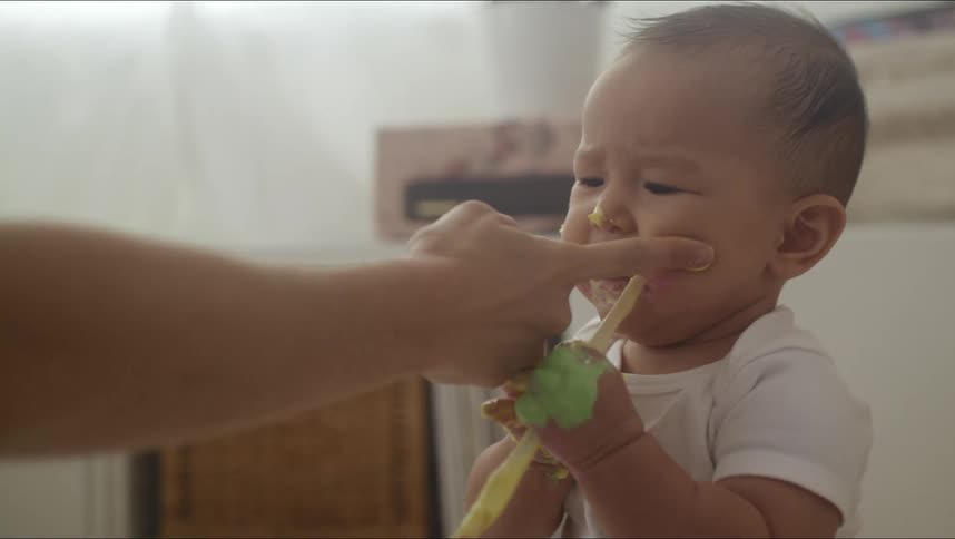 Baby holds a toothbrush in its mouth while an adult touches the babys cheeks and nose with a finger