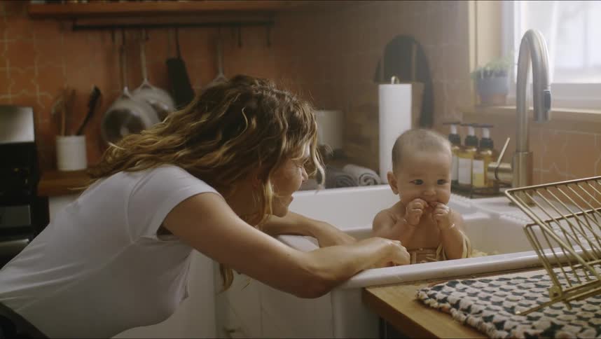 A mother watches her infant play with pasta in a kitchen sink