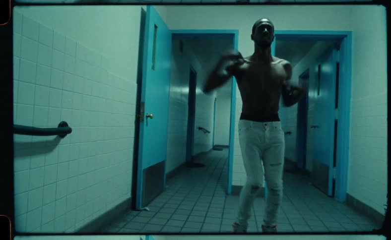 Shirtless black man dancing in an empty corridor with blue doors behind him
