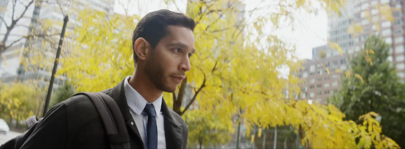 Man wearing a shirt and tie carries something over his shoulder as he walks near the skyscrapers in the city