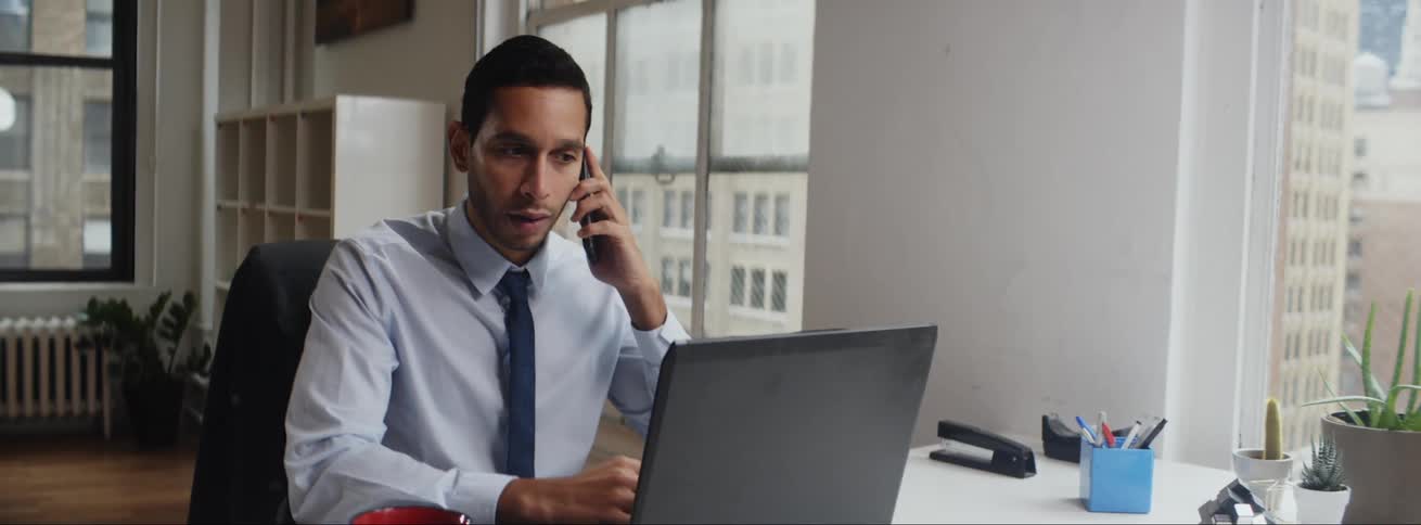 A man is sitting at a desk in an office typing on a laptop computer and he answers his phone and talks to someone