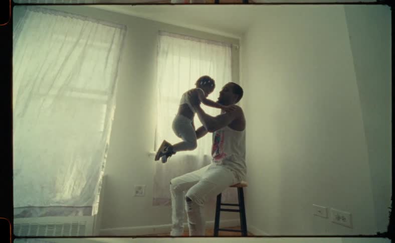 Father wearing white sleeveless shirt and white ripped jeans holds and lifts up daughter sitting on stool in room next to windows